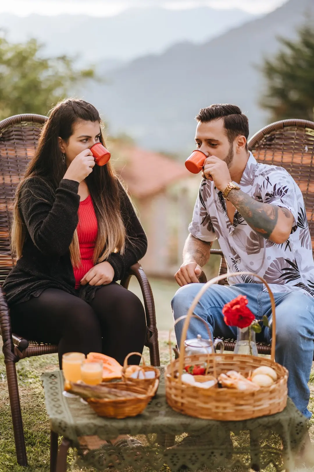 Casal tomando café em canecas esmaltadas nas cadeiras do jardim, com as montanhas ao fundo