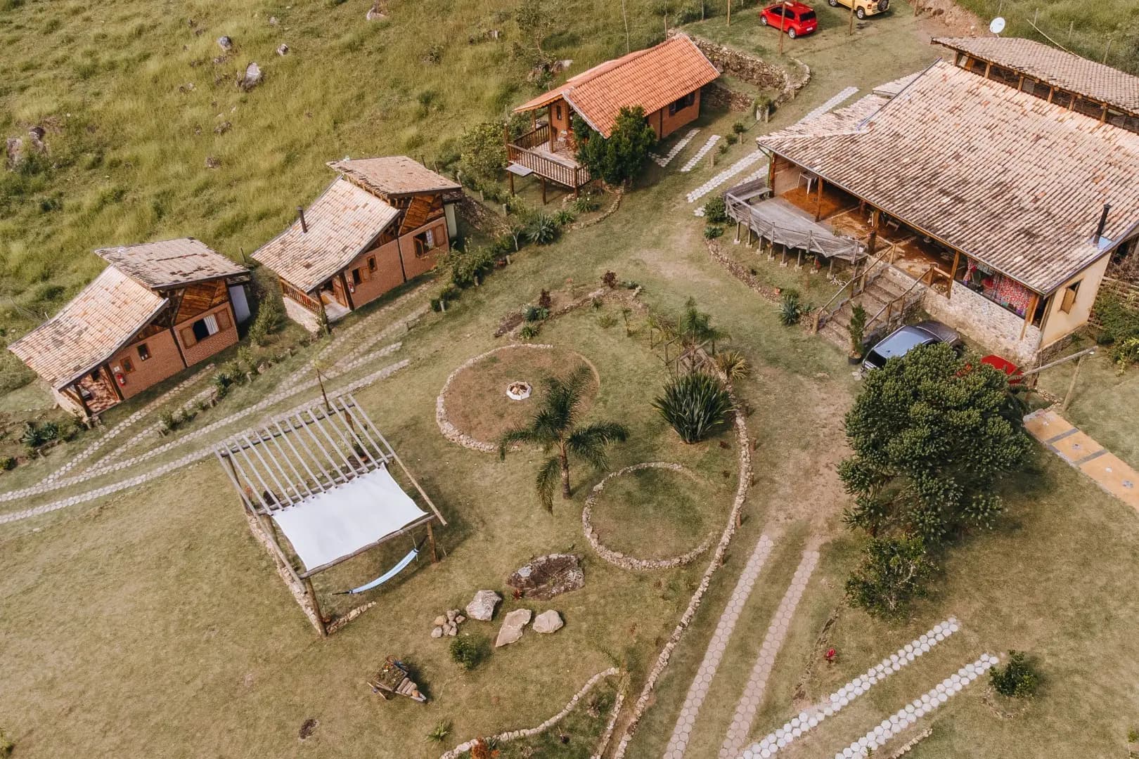 Vista aérea da pousada: chalés, pergolado com redes, roda de fogueira e a casa principal no gramado